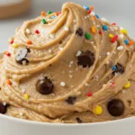 Creamy protein cookie dough with dark chocolate chips, rainbow sprinkles, and sea salt flakes in a white bowl. Close-up of healthy, no-bake edible cookie dough recipe on a minimalist kitchen counter.